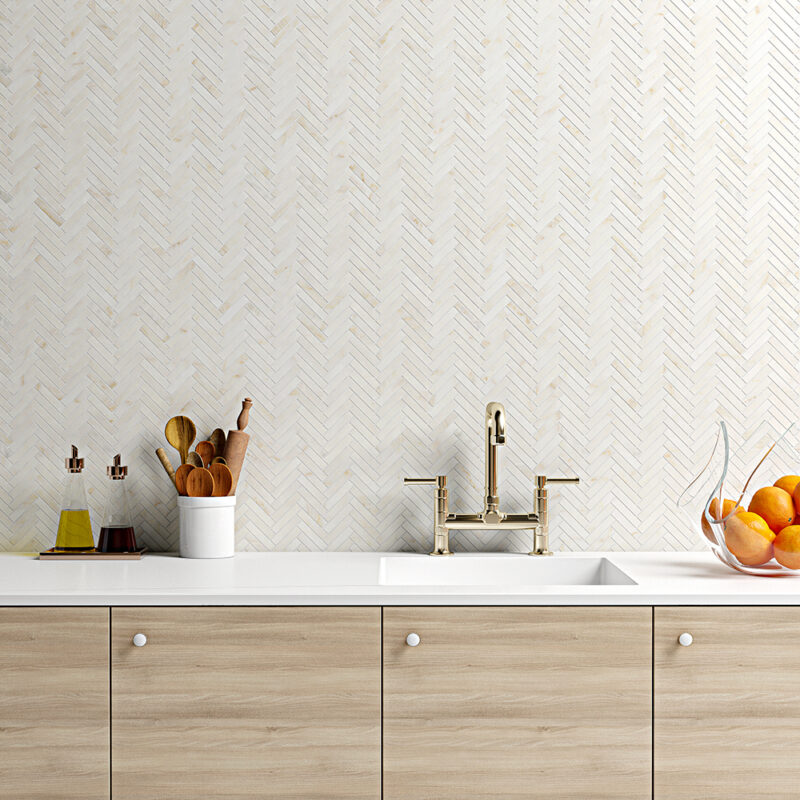Contemporary kitchen with beige herringbone accent wall tile backsplash, paired with light wood cabinets, white countertops, and warm metallic fixtures.